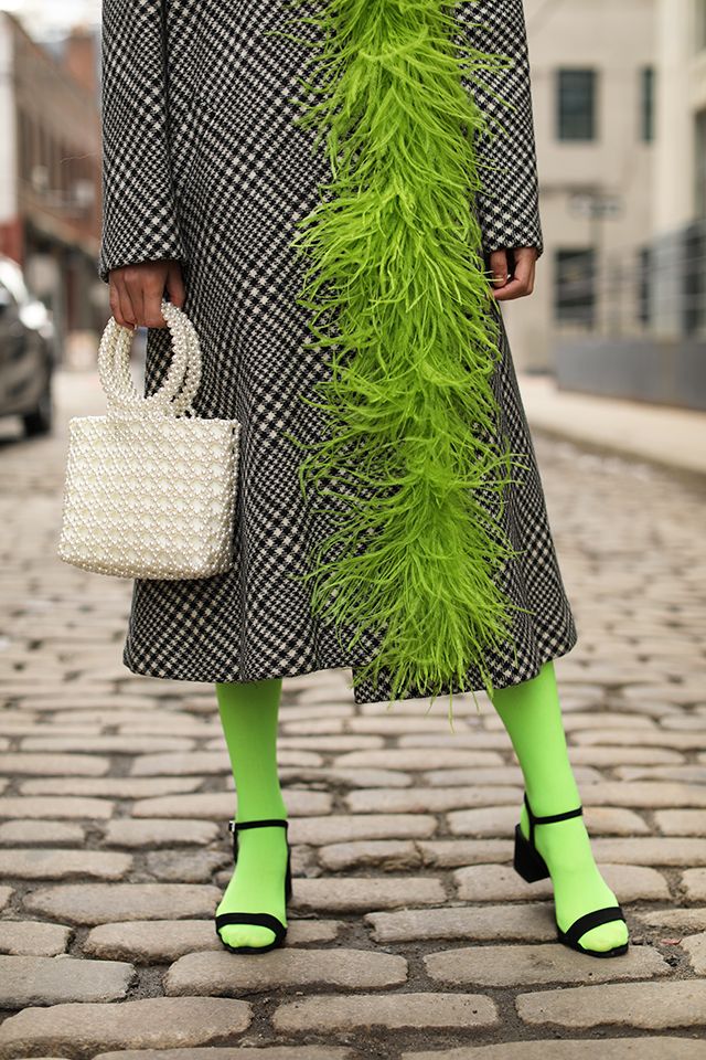 a woman strolls down a cobblestone street wearing a stylish black and white checkered coat adorned with a vibrant lime green feather trim.  she's paired the coat with bright lime green tights and black anklestrap sandals.  she carries a white pearl beaded handbag. the overall style is a chic and eyecatching blend of textures and colors, perfect for autumn or winter fashion.
