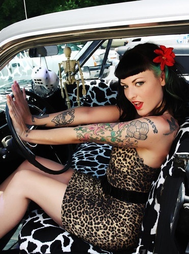 a young woman with extensive tattoos sits in a classic car, wearing a leopard print dress and a red flower in her hair. her style is reminiscent of a rockabilly pinup girl from the 1950s.  she has prominent bangs and vibrant tattoos on her arms. the car's interior has a cowprint seat cover. a skeleton and dice hang from the rearview mirror.