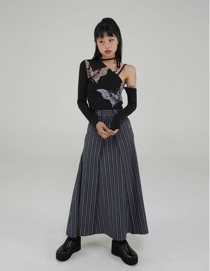a fulllength studio shot of a young asian woman modeling a stylish outfit. she's wearing a black longsleeved top with a oneshoulder design and a subtle butterfly print. the top is paired with a dark grey pinstripe maxi skirt that has a pleated, aline silhouette. she accessorizes with a simple black choker necklace and black platform clogs. the model is posing against a neutral light grey background.