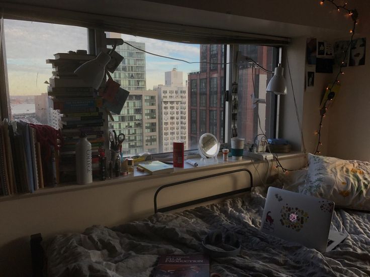 the image shows a dorm room or small bedroom with a large window overlooking a cityscape.  the windowsill is cluttered with books, personal items, lamps, and a few decorative pieces. a bed sits in front of the window, partially covered with a grey blanket. a laptop is on the bed.  the overall atmosphere is cozy and somewhat cluttered, with a mix of organized and unorganized items. fairy lights add a warm glow to the room. the scene suggests a student's living space. the time of day appears to be late afternoon or early evening, judging by the light.
