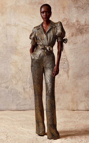 a fulllength studio shot of a darkskinned female model wearing a gold metallic jumpsuit. the jumpsuit features a collared shirtstyle top with puff sleeves and a wideleg, flared bottom.  the fabric has a geometric patterned texture. the model is standing against a neutral beige background.  the overall style is sophisticated and elegant.