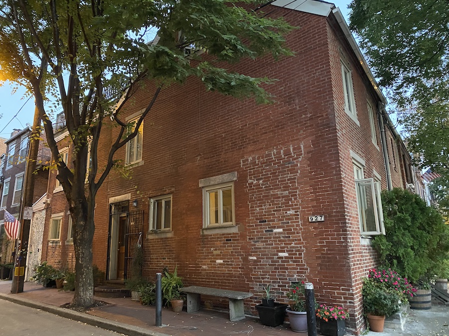 a brick building with several windows, a bench, plants, and flowers on a street. an american flag is visible in the distance. the building has the number 927 on its exterior. a tree is visible in the foreground.