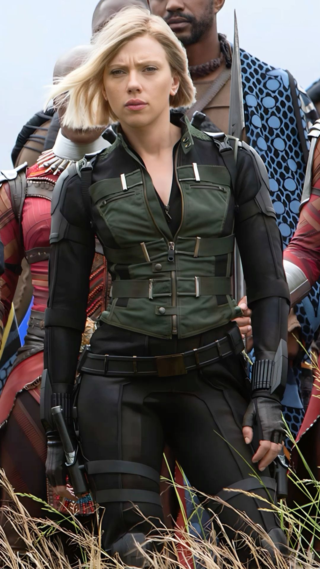 a closeup of black widow, portrayed by scarlett johansson, in her black and green costume. she is standing in a field of tall grass, looking intently at the camera. she is wearing a black leather jacket over a black shirt, black pants, and black boots. her hair is styled in a bob and she is wearing a black belt. she is holding a gun in her right hand and her left hand is resting on her hip. she is looking straight ahead with a serious expression on her face.