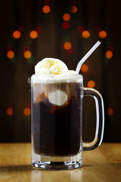 a closeup shot of a root beer float, a classic and delicious dessert drink, served in a glass mug with a straw. the ice cream is melting on top of the dark soda, creating a beautiful and tempting visual.  the image is taken against a blurry background of warm, festive lights, likely suggesting a bar or restaurant setting.