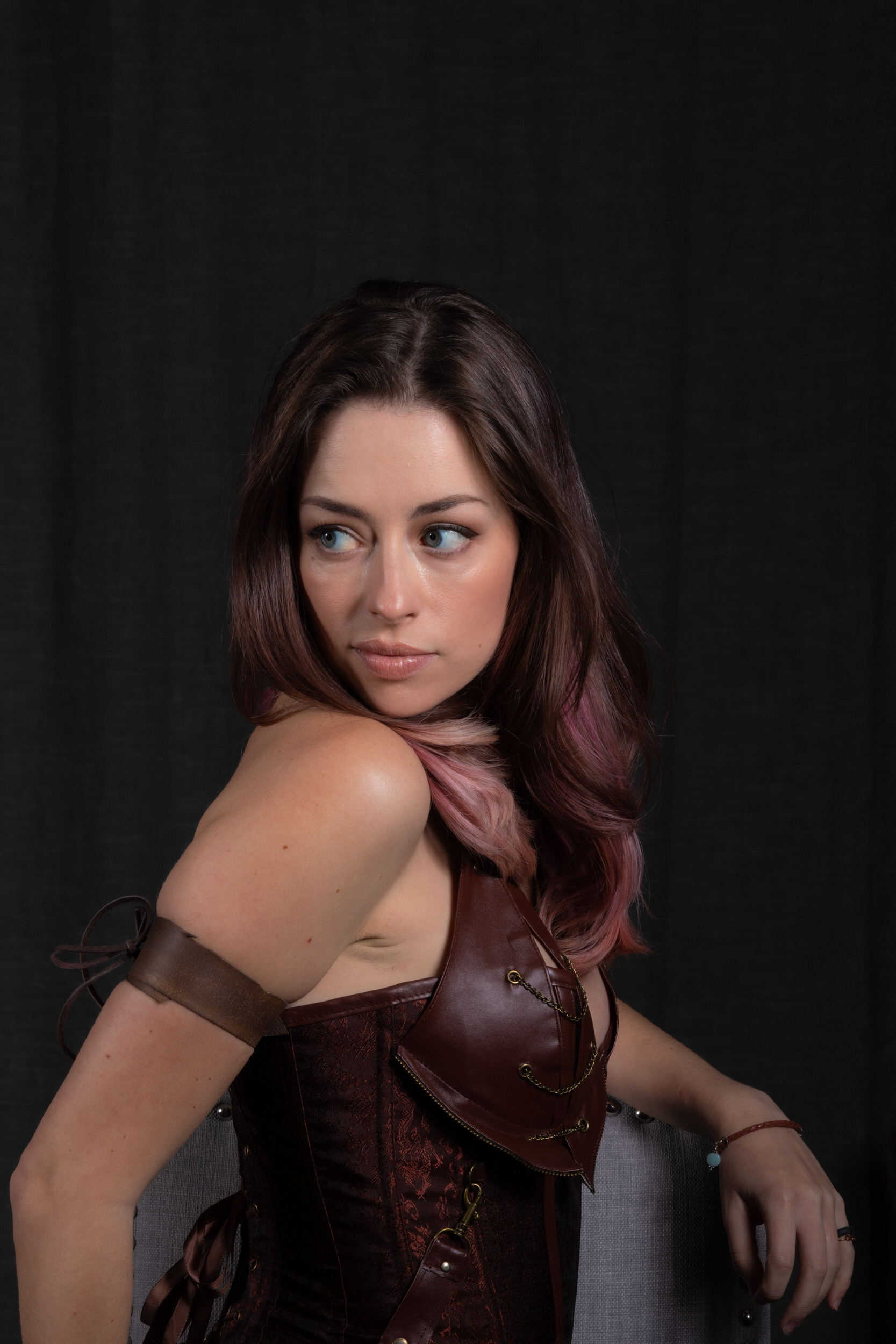 a woman with long brown hair streaked with pink is posed against a black backdrop. she is wearing a brown leatherlook corset top with metal accents and a brown leatherlook bracer on her right arm. she is leaning on a grey stool and looking over her shoulder to the left of the frame. the image appears to be lit with artificial light.