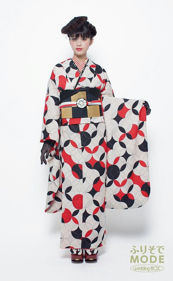 a young woman is standing and facing forward in a brightly lit studio setting, wearing a traditional japanese kimono with a bold geometric pattern. the kimono features a combination of offwhite, red, and black circular and diamond shapes. she has a wide black obi (sash) with gold and white accents tied around her waist. her hair is styled in a distinctive updo with a small, dark headpiece. the woman has fair skin and a neutral expression. her right hand is visible and covered with a black lace glove. the background is plain white, and the photo is welllit, highlighting the textures and colors of the kimono. in the lower right corner, there's a small logo or text in japanese.