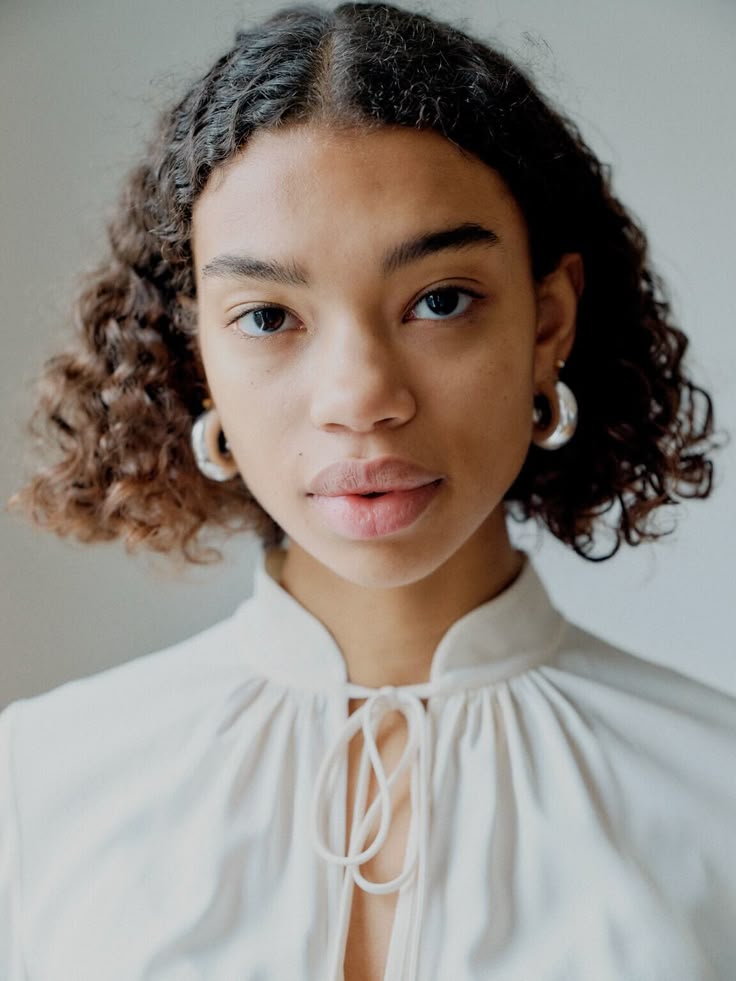 a head and shoulders portrait of a young woman with curly dark hair, wearing silver hoop earrings and a white blouse with a tie neck detail. she is looking directly at the camera.