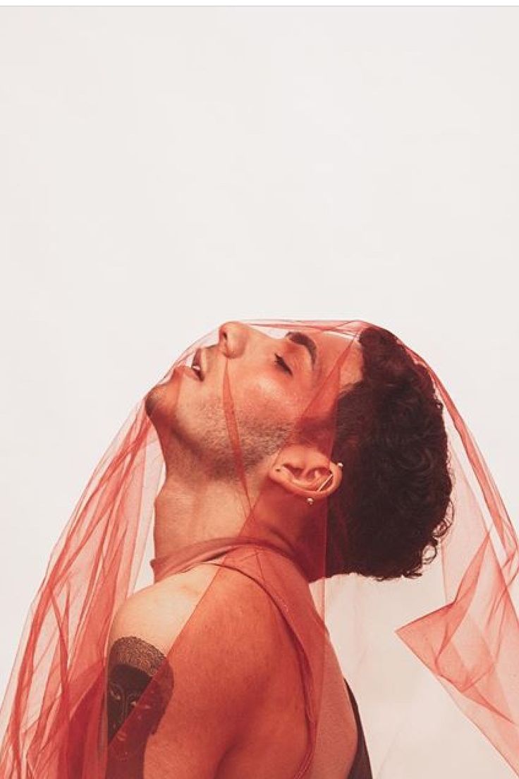 a closeup portrait of a man with his eyes closed and head tilted back, shrouded in a delicate red veil. his skin is soft and smooth, and his hair is curly and dark.  he is wearing a small hoop earring in his ear, and his chest is bare.  he has a tattoo on his shoulder.  the image is set against a white background.