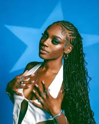 a young black woman with long black braids, wearing a white sleeveless top and silver jewelry, poses against a blue backdrop. she is looking at the camera and has a confident expression. her makeup is subtle with a nude lip and dark eyeshadow. she looks edgy and stylish.