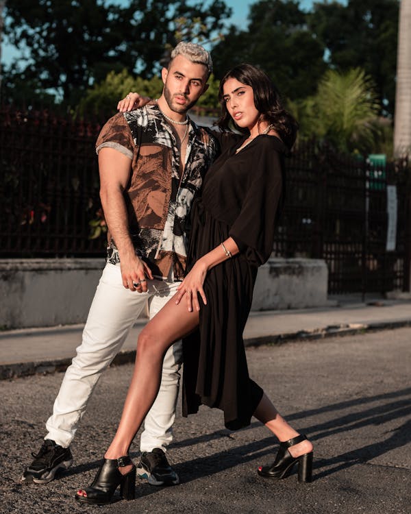 the image shows a stylish young couple posing on a city street. the man is wearing white pants and a printed shortsleeved shirt, while the woman wears a black dress and high heels. they are standing close together, with the woman's arm around the man's shoulders, showcasing an affectionate pose. the setting appears to be an urban environment in the late afternoon, with warm sunlight casting shadows on the street. the overall style is trendy and fashionforward, suggesting a photoshoot or street style photography. the couple looks confident and comfortable, highlighting their coordinated outfits and stylish look.