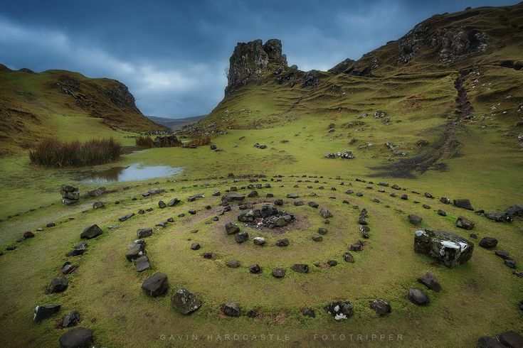 this is a landscape photograph of a valley surrounded by green hills under a cloudy sky. in the foreground, there is a stone spiral or circle made of rocks on a grassy area. in the background, there is a pond and a rock formation. the photo was taken in the isle of skye, scotland, possibly in the quiraing or fairy glen area.