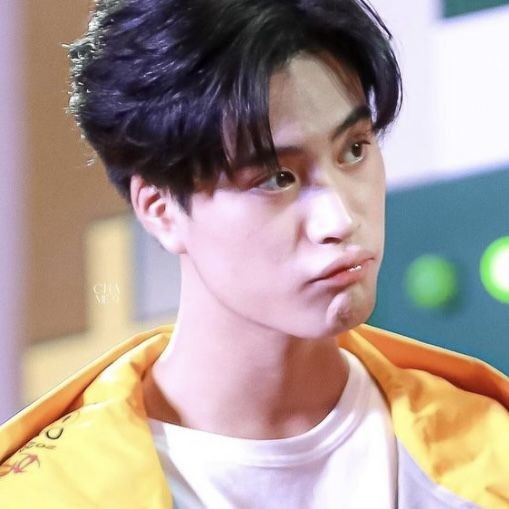 a closeup portrait of a young asian man with black hair styled with bangs and a side part. he is wearing a yellow jacket and a white shirt. he has a slightly pouty expression on his face. the background is blurred, and the lighting is bright and artificial.