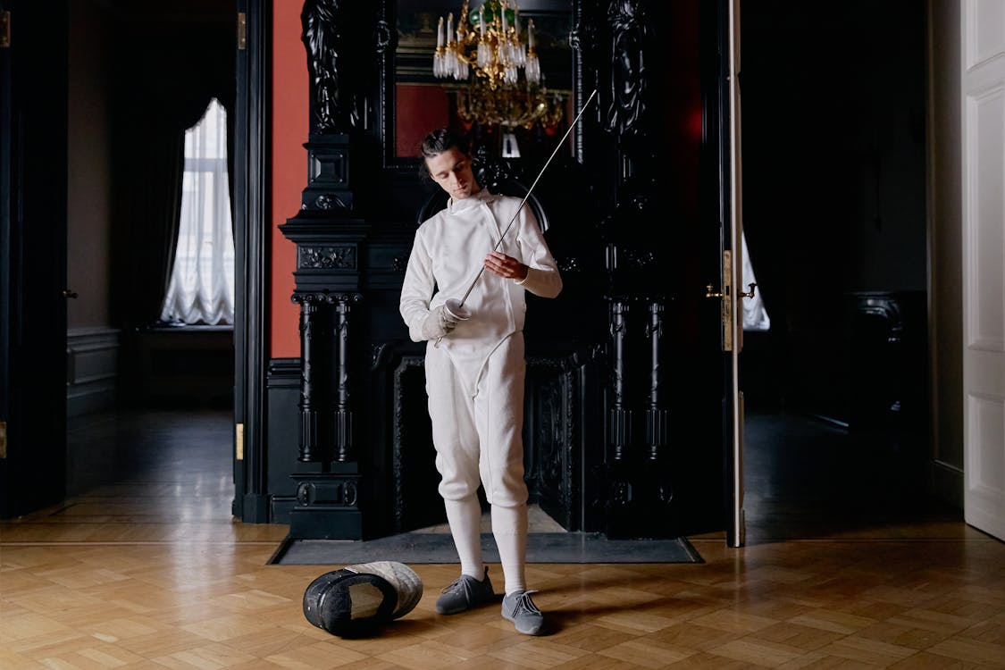 a fulllength shot of a male fencer in a white uniform standing in an elegant, vintage interior. he is holding a foil and appears to be inspecting or preparing his equipment. a fencing mask rests on the floor beside him. the setting is grand, with a dark wood fireplace and ornate details in the background. the image is both dramatic and stylish, creating a sense of classic athleticism.