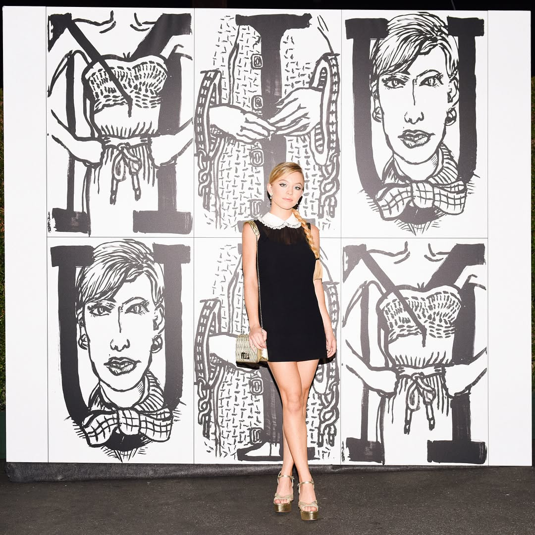 a young blonde woman in a black dress with a white peter pan collar stands in front of a wall featuring a repeating black and white graphic design. the design appears to be handdrawn and features stylized female figures in various poses. the woman is carrying a small handbag and is wearing gold highheeled shoes. she looks directly at the camera. the overall style of the image is fashionable and chic, with a blend of high fashion and artistic elements. the image could be a promotional shot from a fashion event or a publication showcasing a designer's work.