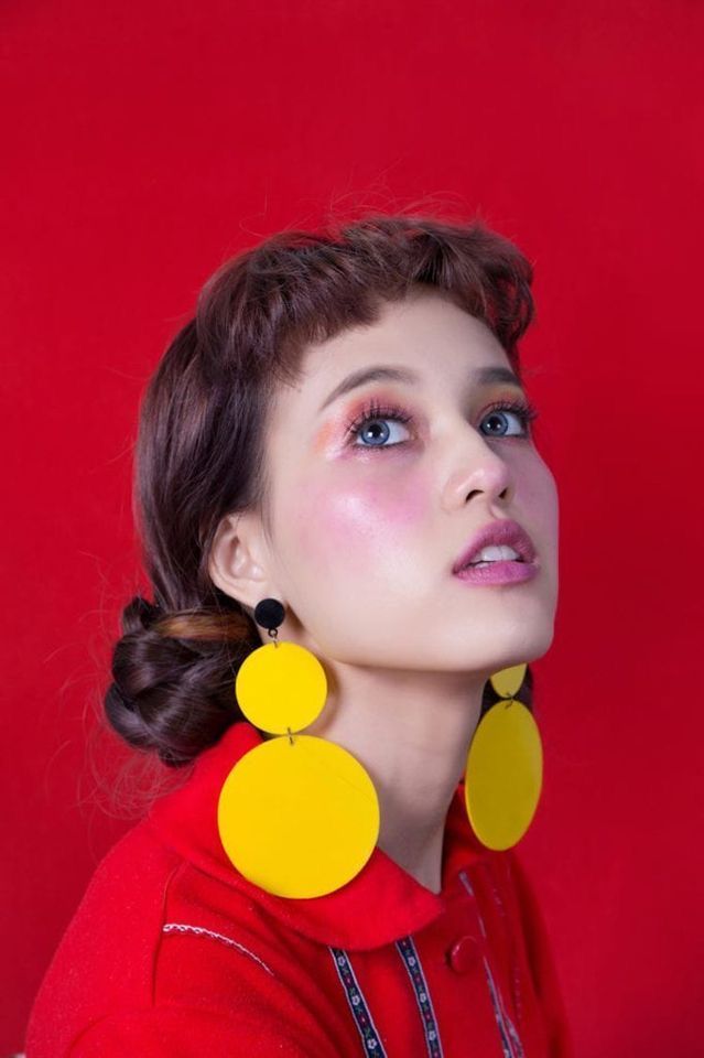 loseup portrait of a young woman with dark brown hair styled in a bun. she is wearing a red collared shirt and large, statement yellow circular earrings. her makeup is noticeable, with blush and eye makeup.  the background is a solid, vibrant red. the overall style is bold and retroinspired, with a strong contrast between the red and yellow colors.  the woman's expression is one of looking upward.