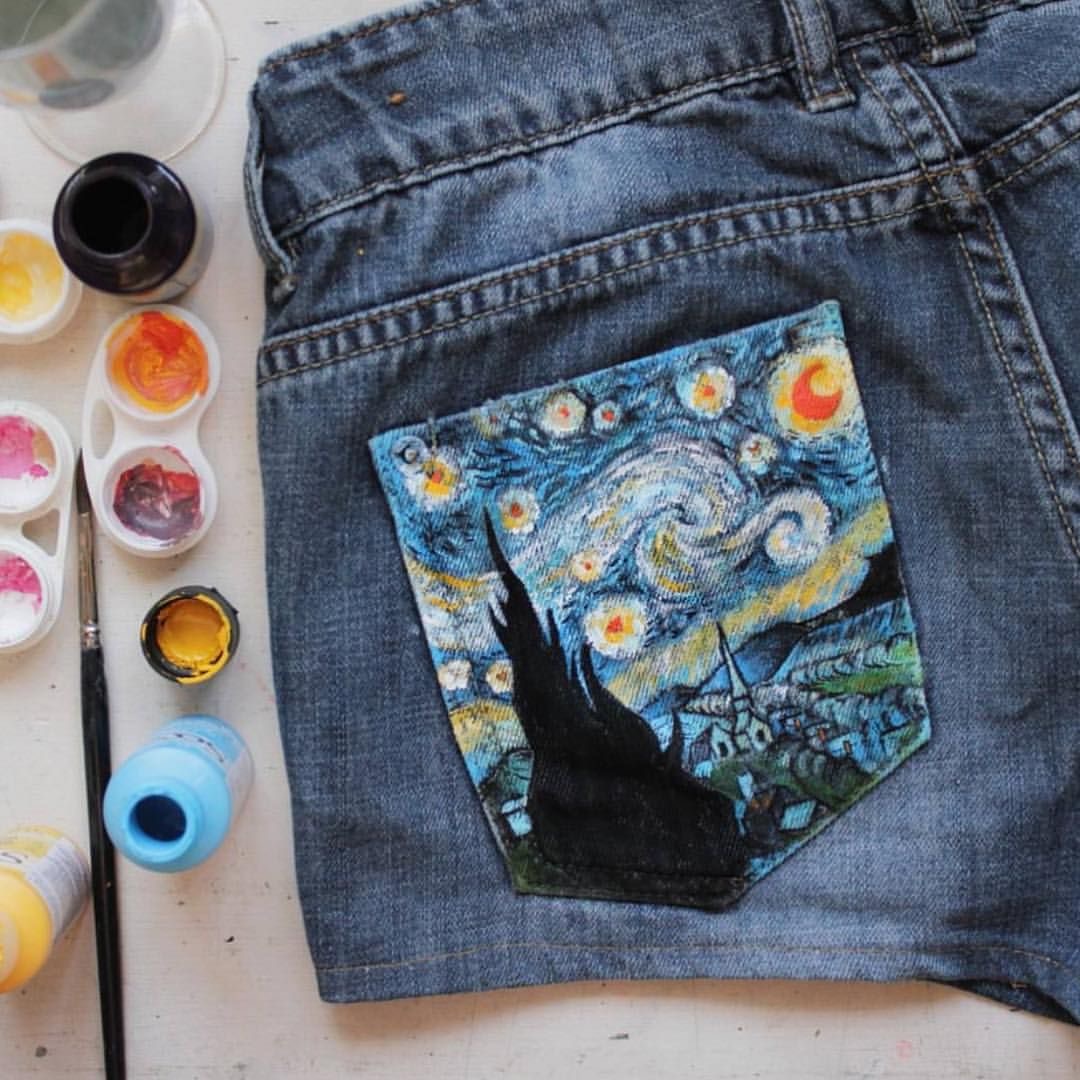 a pair of denim shorts lying on a white surface. the back pocket of the shorts has a painting of van gogh's starry night. there are paint bottles and an opened paint palette with brushes next to the shorts.