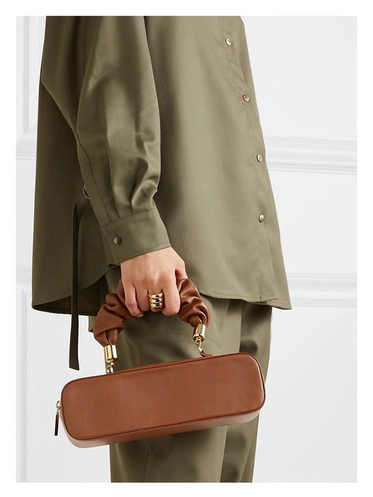 the image shows a closeup of a woman's hand holding a brown leather boxshaped handbag. the bag features a unique gathered fabric top handle with gold hardware accents. the woman is wearing a loosefitting, olivegreen shirt.  the overall style suggests a highend, minimalist fashion aesthetic. the focus is clearly on the handbag, highlighting its design and texture.