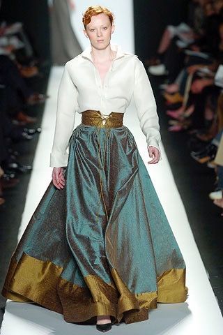 a model walks the runway during a fashion show. she is wearing a longsleeved, white buttondown shirt, a highwaisted, full green skirt with a gold hem, and a wide gold belt. the runway is white, and there are people in the audience on either side.