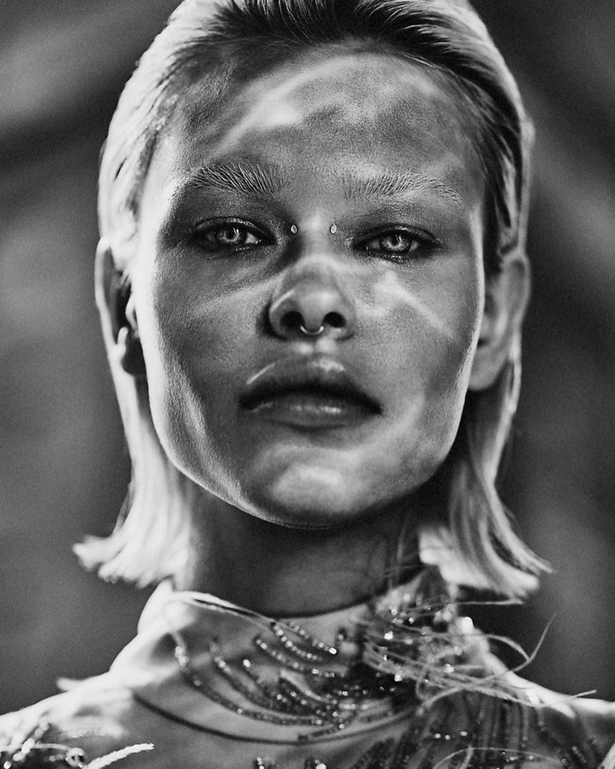 a black and white portrait of a woman with short blonde hair. she is looking directly at the camera with a serious expression. she is wearing a beaded top and a nose ring. the image is closeup and the focus is on her face. the lighting is dramatic, creating strong shadows and highlights. the overall effect is intense and striking.