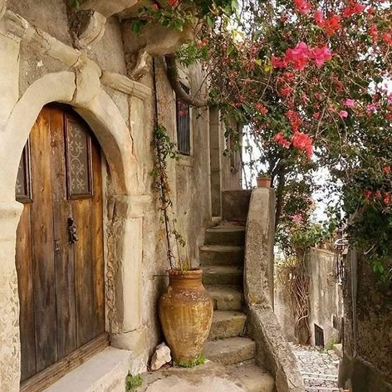 the image shows a weathered stone building with an arched wooden door. to the right of the door are stone stairs leading upwards, adorned with a large vase and lush bougainvillea flowers. the setting appears to be an old alleyway.