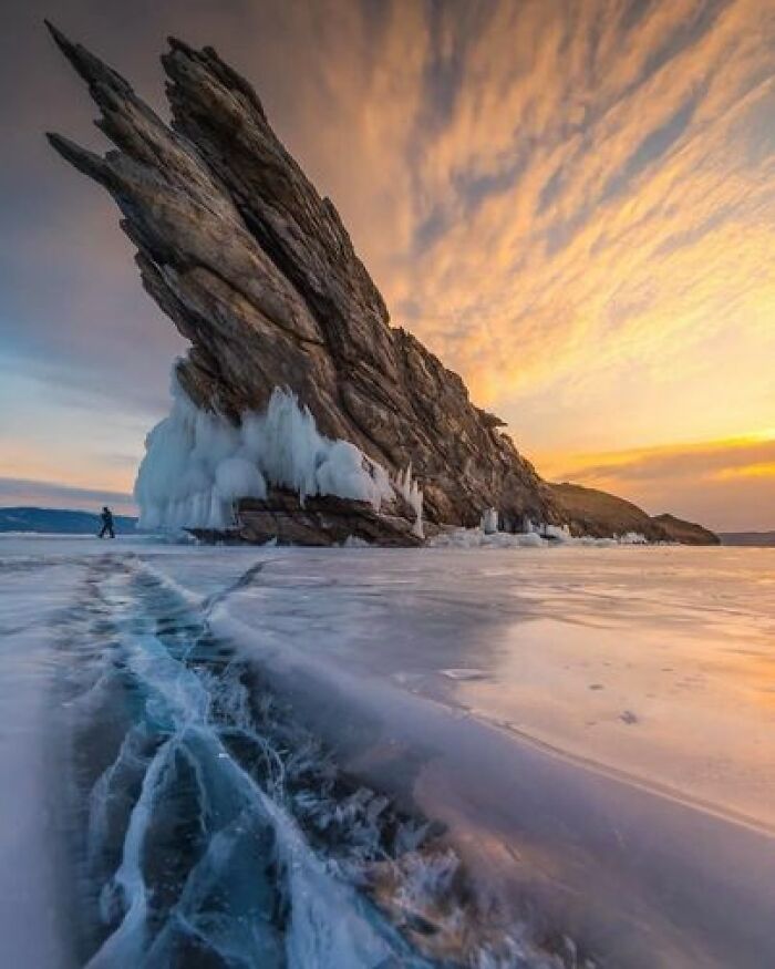 a stunning landscape photograph shows a large, jagged rock formation covered with icicles rising dramatically from a frozen lake under a sunset sky. a prominent crack runs through the ice in the foreground, leading towards the rock. a person is walking in the distance near the rock. the sky is filled with colorful clouds.