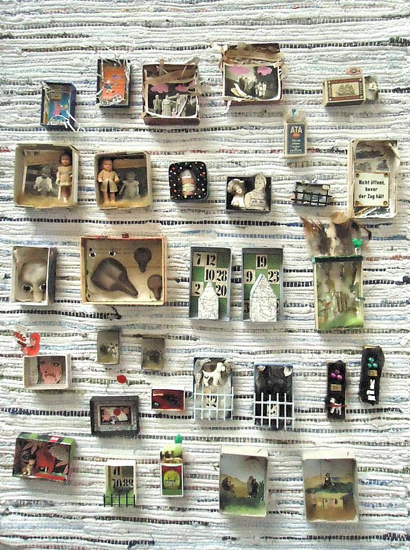 the image shows a collection of miniature scenes and objects displayed within small, possibly repurposed matchboxes. these boxes are arranged in a gridlike fashion on a woven rug or textile with a striped pattern. the contents of the boxes range from figurines and small objects to pictures and fragments, creating a collagelike effect. the overall impression is of a handcrafted, vintage collection of mementos and found objects.