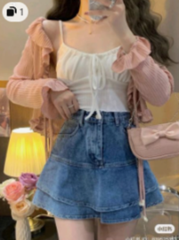 a young woman stands indoors taking a mirror selfie. she is wearing a white top with a tie front detail and a pink ruffled cardigan. she has paired this with a denim mini skirt and is carrying a pink bag with a bow. the background is a bedroom with a lamp and flowers.