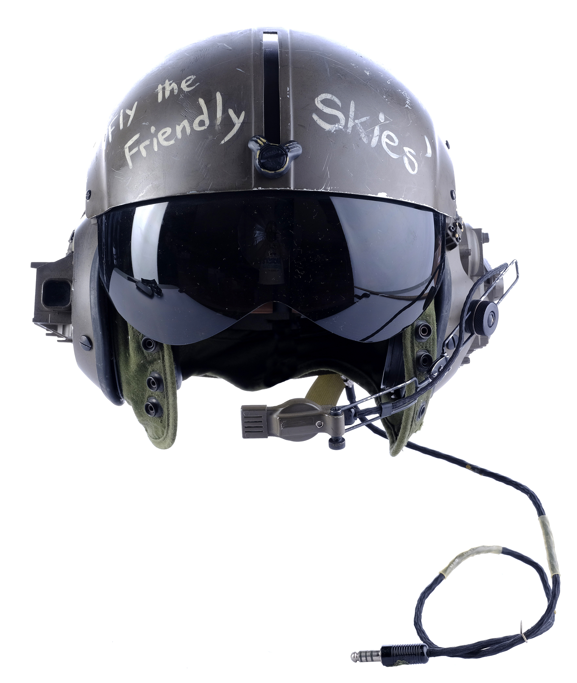 a closeup of a military flight helmet. the helmet is painted green and has a black visor. the helmet has a microphone attached to it, and there is a cord extending from the helmet.  there is a handwritten message on the side of the helmet that says "fly the friendly skies"  in white paint.