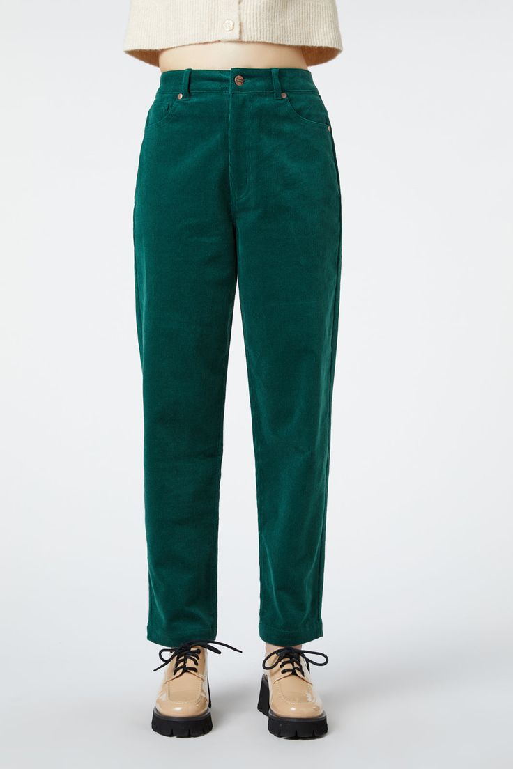 a woman is wearing a pair of highwaisted, relaxedfit green corduroy pants. she is standing in front of a white background and is wearing tan laceup leather shoes. the woman is looking at the camera.