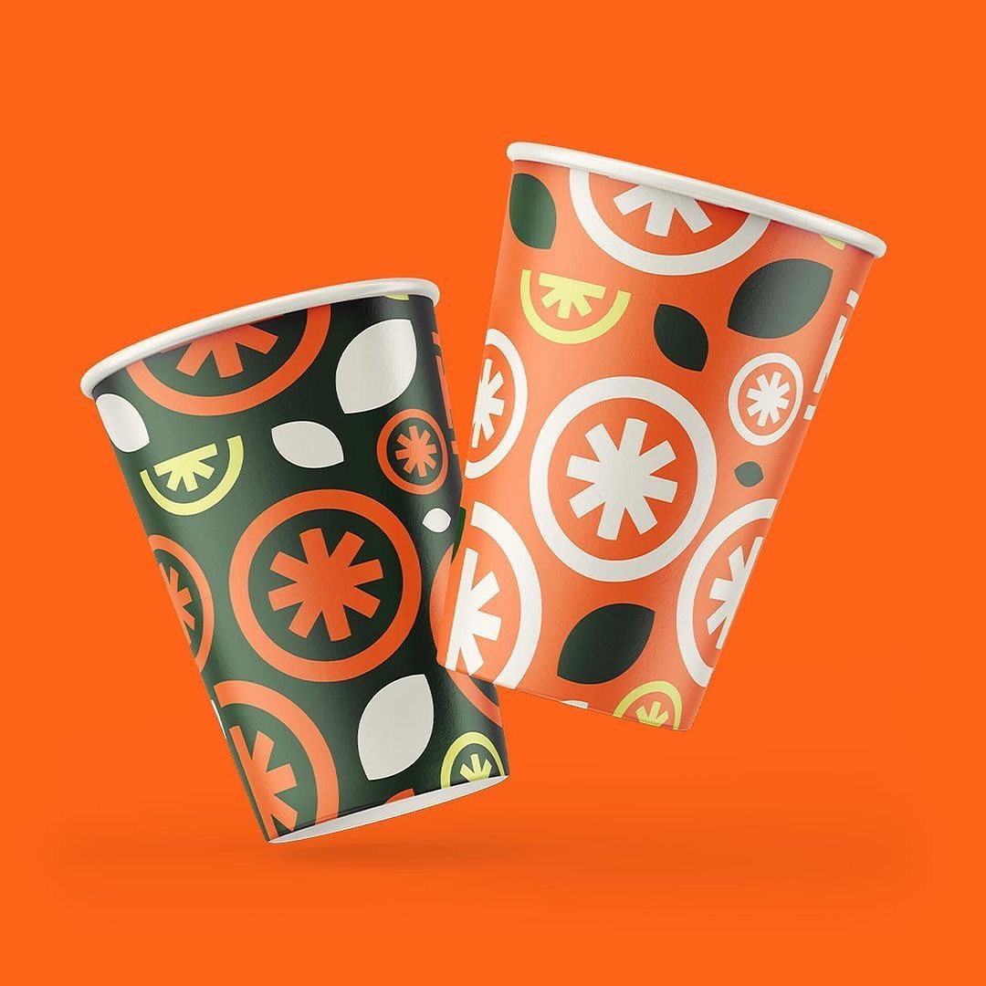 the image shows two disposable paper cups with a vibrant and modern design. the cups are placed against a bright orange background. the design on the cups features a repeating pattern of stylized orange slices, leaves, and abstract floral motifs in a color palette of orange, green, white, and dark gray. the cups are presented at a slight angle, with one cup slightly tilted forward, as if in motion, adding a dynamic element to the composition.