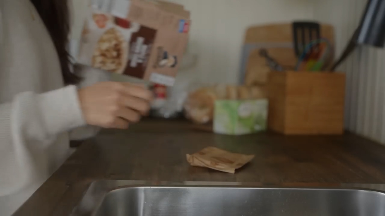 a woman stands in a softly lit kitchen, holding a box of cereal. she appears to be in the process of preparing breakfast, with her focus on the task at hand. the warm, neutral tones of the kitchen create a cozy and inviting atmosphere.