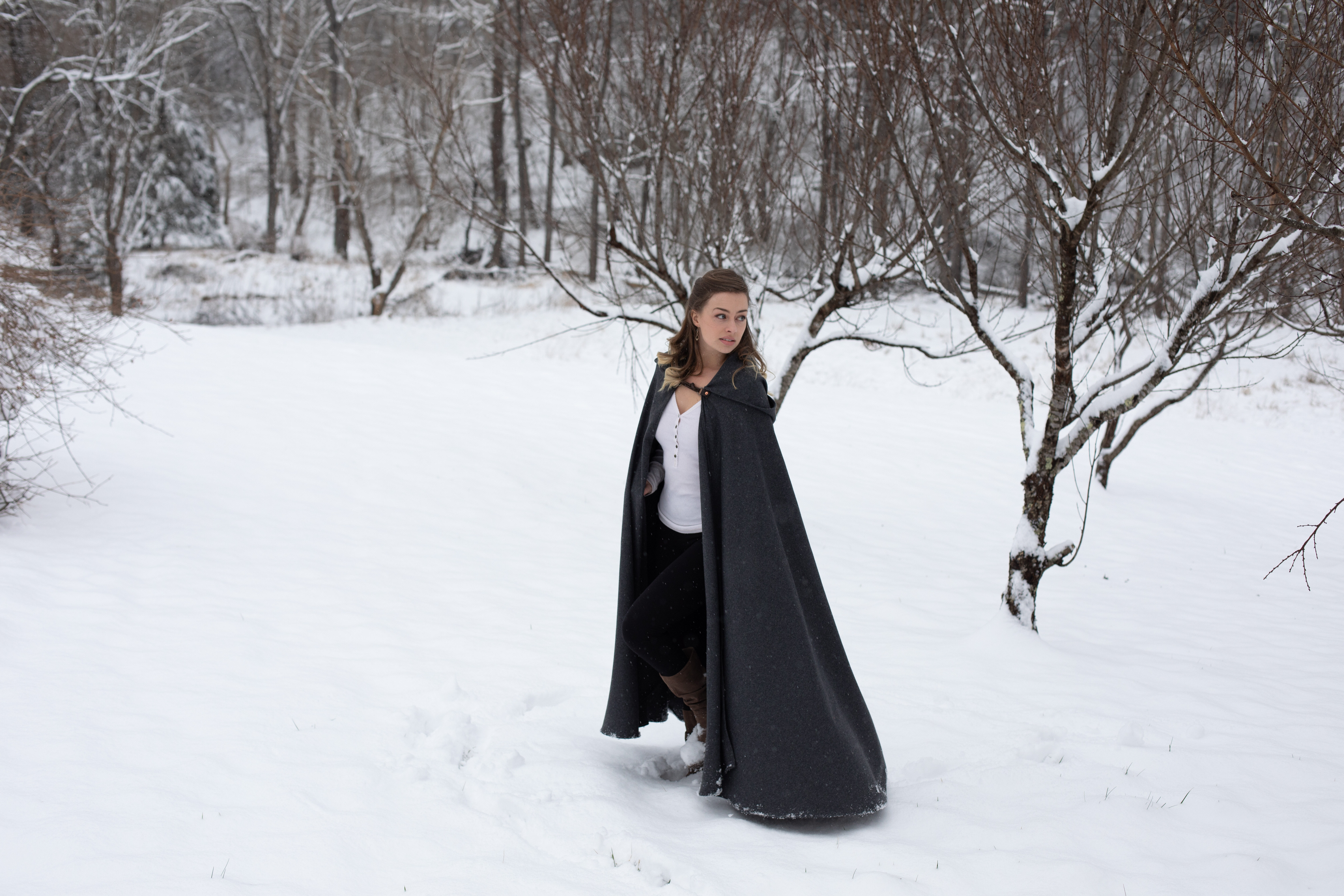 a young woman in a long, dark gray hooded cape walks through a snowy forest. she is wearing black pants and brown boots, and her hair is pulled back in a loose braid. the trees are bare and covered in snow, and the ground is blanketed in white. the overall feel of the image is one of peace and tranquility.