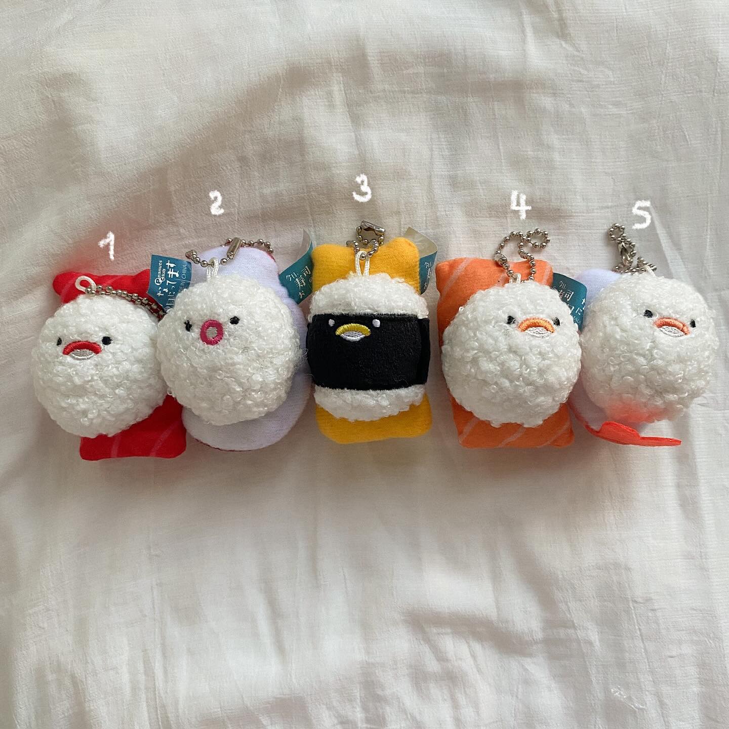 a line of five plush sushi keychains with different types of sushi, each topped with an embroidered bird character, set against a white sheet. from left to right, there is a tuna sushi with a simple smile, an octopus sushi with a small round mouth, an egg sushi with a penguin face, a salmon sushi with an orange beak, and another octopus sushi with an open mouth. all of the keychains have silver chains.