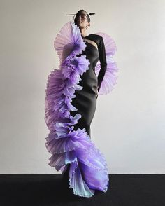 a woman poses in a black dress with a purple and light blue tulle overlay that has a ruffled, flowing look. the dress is tightfitting in the torso and waist and flows out in a long, elegant skirt. the woman's hair is dark and she is wearing a dark eye makeup look.  she's standing on a black and white backdrop.