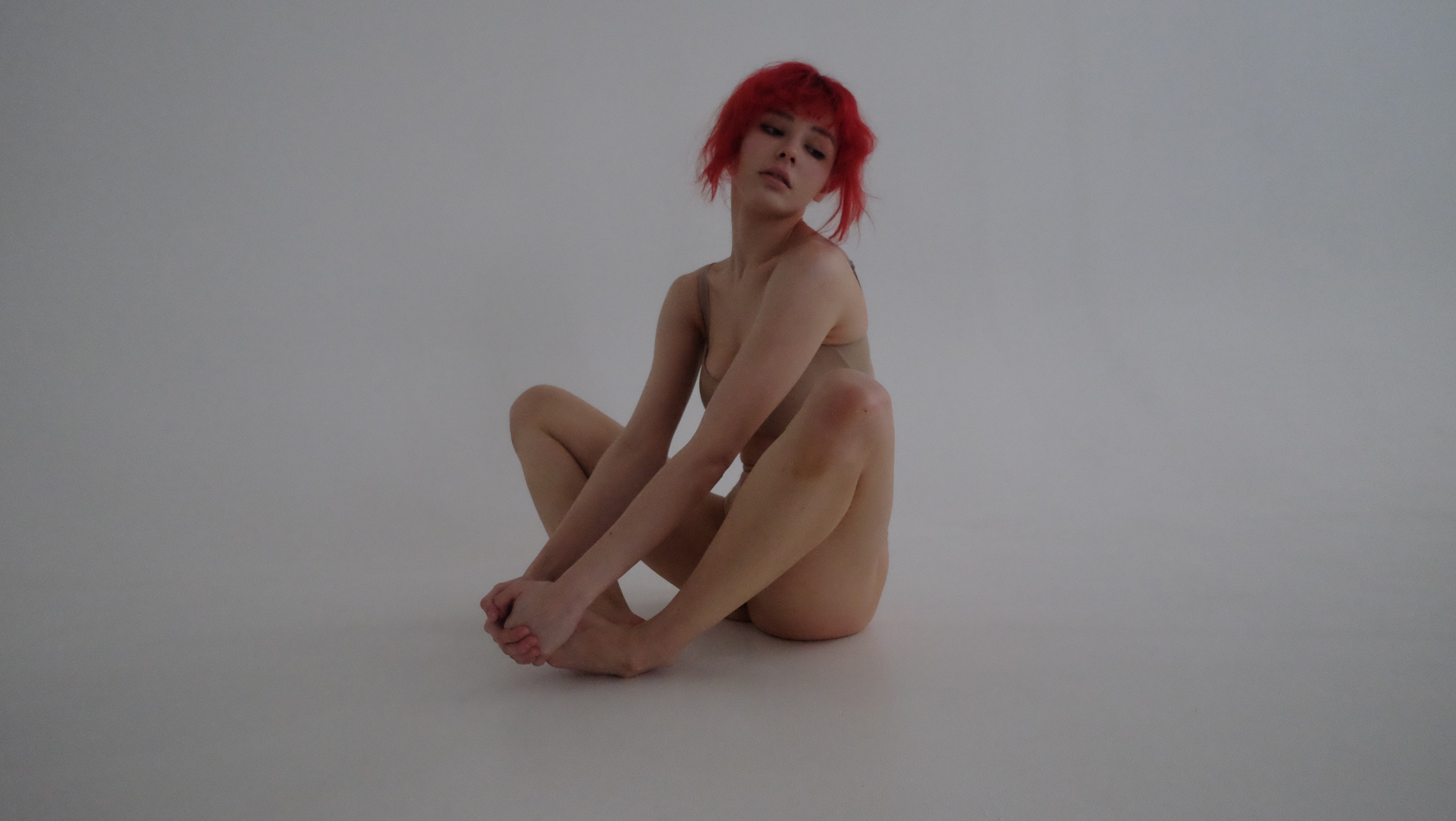 a woman with short red hair is sitting on a white studio floor in a dancer's pose, her legs are crossed and her arms wrapped around them. she is wearing a nudecolored bodysuit and looking directly at the camera. the image has soft, natural lighting.