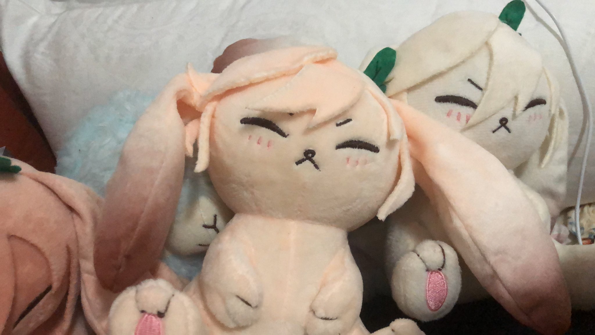 three plush bunny toys are sitting on a white fluffy blanket. the bunny in the center is pink and white, with big brown eyes, a pink nose, and a brown mouth. it has long, floppy ears. the bunny on the left is white with brown ears and a blue body. the bunny on the right is white with green leaves on its head and brown eyes. all three bunnies are looking at the camera with a cute expression.  the image is full of warmth and happiness.  it's a perfect image to bring a smile to someone's face.