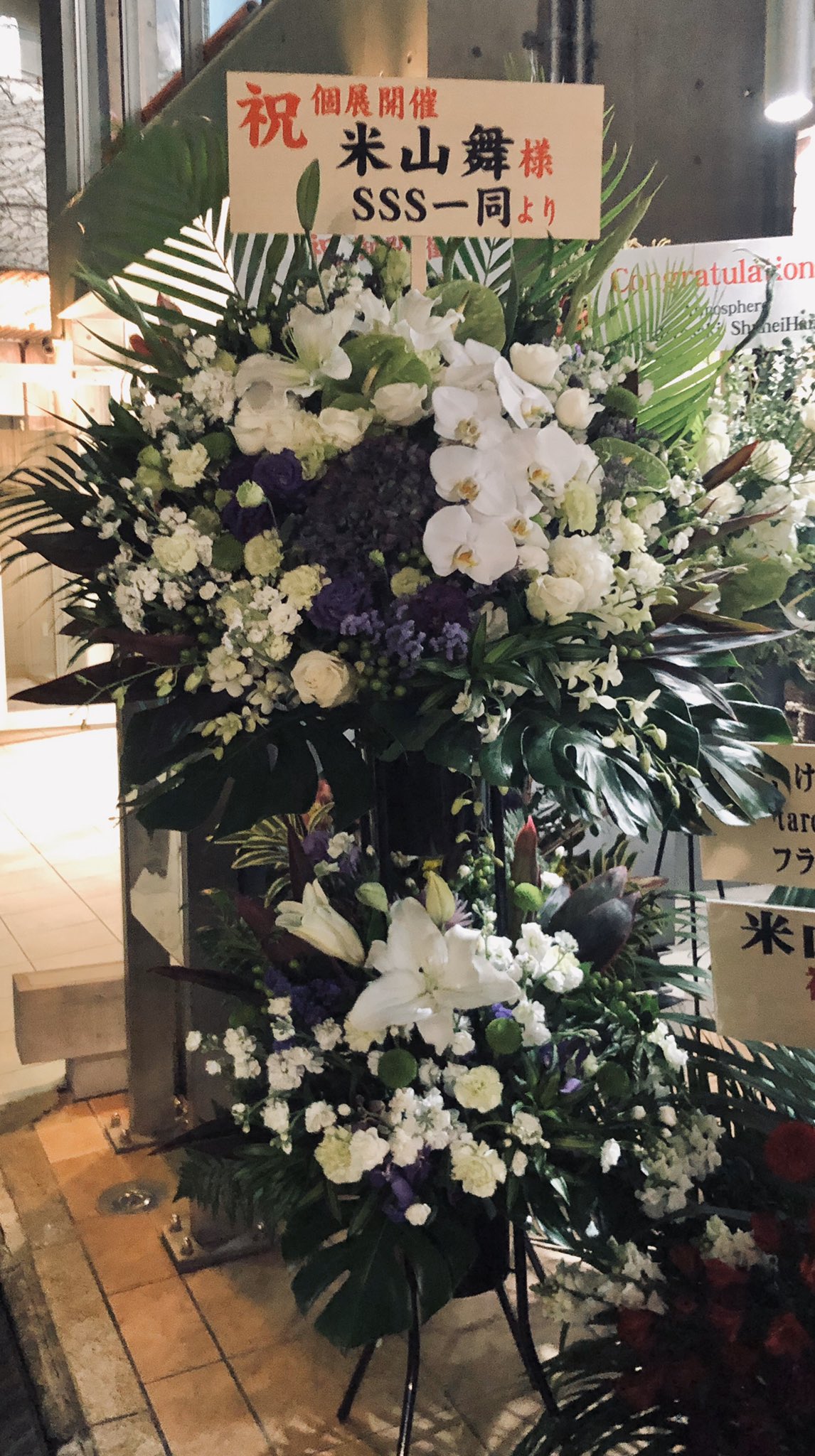 a large, colorful flower arrangement stands in front of a business. the arrangement is primarily white lilies and orchids with accents of purple and green foliage. a sign with japanese characters congratulates someone named "yamamai" on their solo exhibition.  the sign also says "from everyone at sss." the image is taken indoors at the entrance of the business.