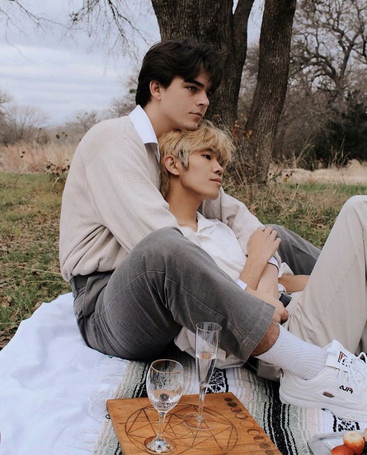 two men are sitting on a blanket in a park. they are holding each other and looking at each other with affection. the man on the left is wearing a cream sweater and grey pants. the man on the right is wearing a white shirt and beige pants. there are two glasses of champagne and a wooden tray on the blanket. the men are surrounded by nature. the background of the image includes trees and grass. the men are smiling and seem very happy. the image suggests a romantic relationship.