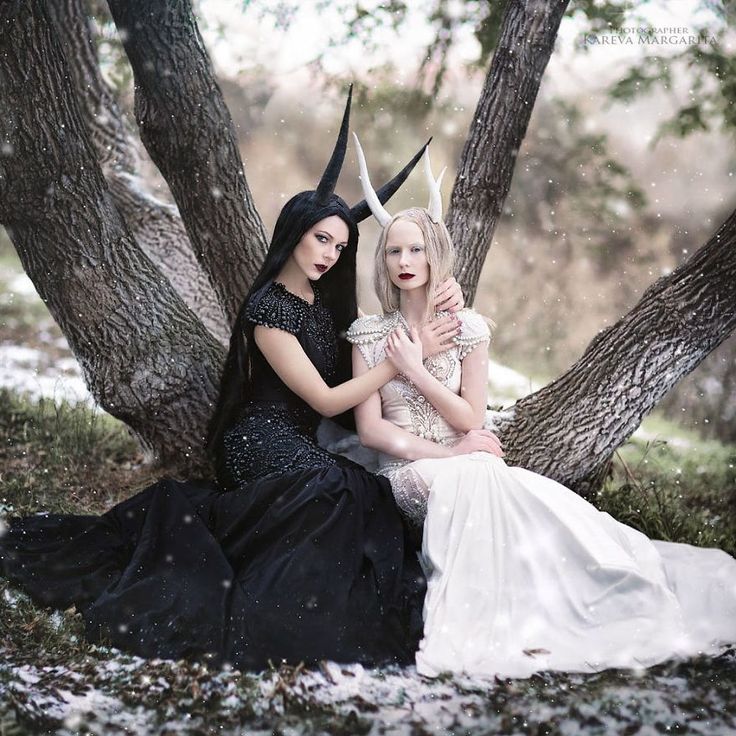 two women, dressed in black and white, sit on a snow covered forest floor. the woman on the left has black horns and is wearing a black dress, while the woman on the right has white horns and is wearing a white dress. they are looking at the camera, with a serious expression on their faces. the forest behind them is covered in snow.