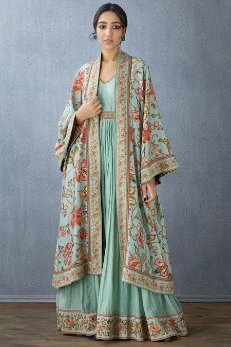 a woman is standing in front of a textured wall, wearing a long, light blue indowestern dress with a floral print shawl. the dress has a gathered or pleated bodice and a long, flowing skirt. the shawl is also light blue with a floral pattern in shades of red, orange, and green, and it features decorative embroidery along the edges. the woman has dark hair pulled back from her face, and she is wearing small earrings. the overall style is elegant and traditional.