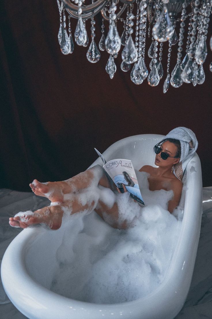 a woman relaxes in a luxurious bathtub filled with bubbles, reading a magazine. she wears sunglasses and a towel on her head. a crystal chandelier hangs above her. the scene is elegant, serene, and peaceful, conveying a feeling of selfcare and indulgence.