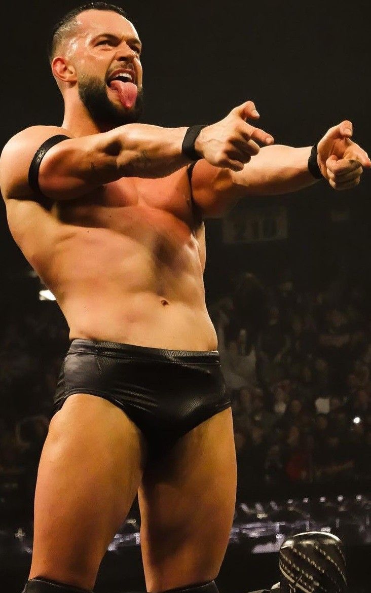 a closeup shot of a muscular male professional wrestler, possibly in the middle of a performance. he's shirtless, wearing black wrestling trunks, and has a confident, cocky expression with his tongue sticking out.  his arms are outstretched, pointing towards the unseen audience in a gesture of showmanship. the background is blurred, suggesting a large audience in a dark arena. the lighting focuses on the wrestler.