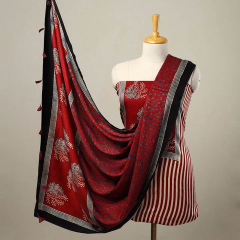 the image shows a mannequin dressed in a red and white striped ethnic wear ensemble. the dress features vertical stripes and a blockprinted floral design on the upper part. a red scarf with a floral pattern and black border is draped over the mannequin. the scarf also has tassels on one end. the outfit appears to be made of cotton fabric.