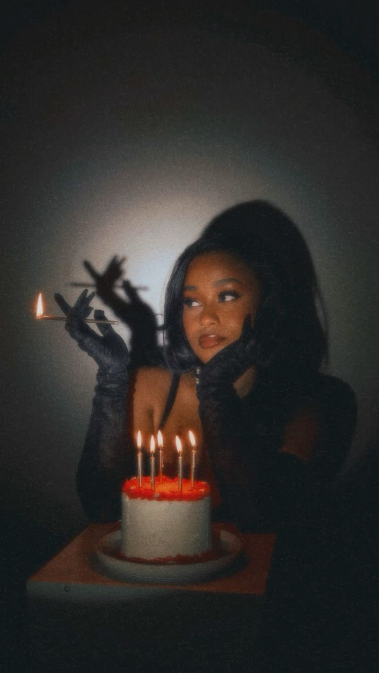 a woman is blowing out the candles on a birthday cake. she is wearing a black dress and gloves, and she is sitting in a dark room. the cake is lit by candles and the woman is smiling. this image captures a moment of joy and celebration, and the dark background adds to the mysterious and elegant feel of the photo.