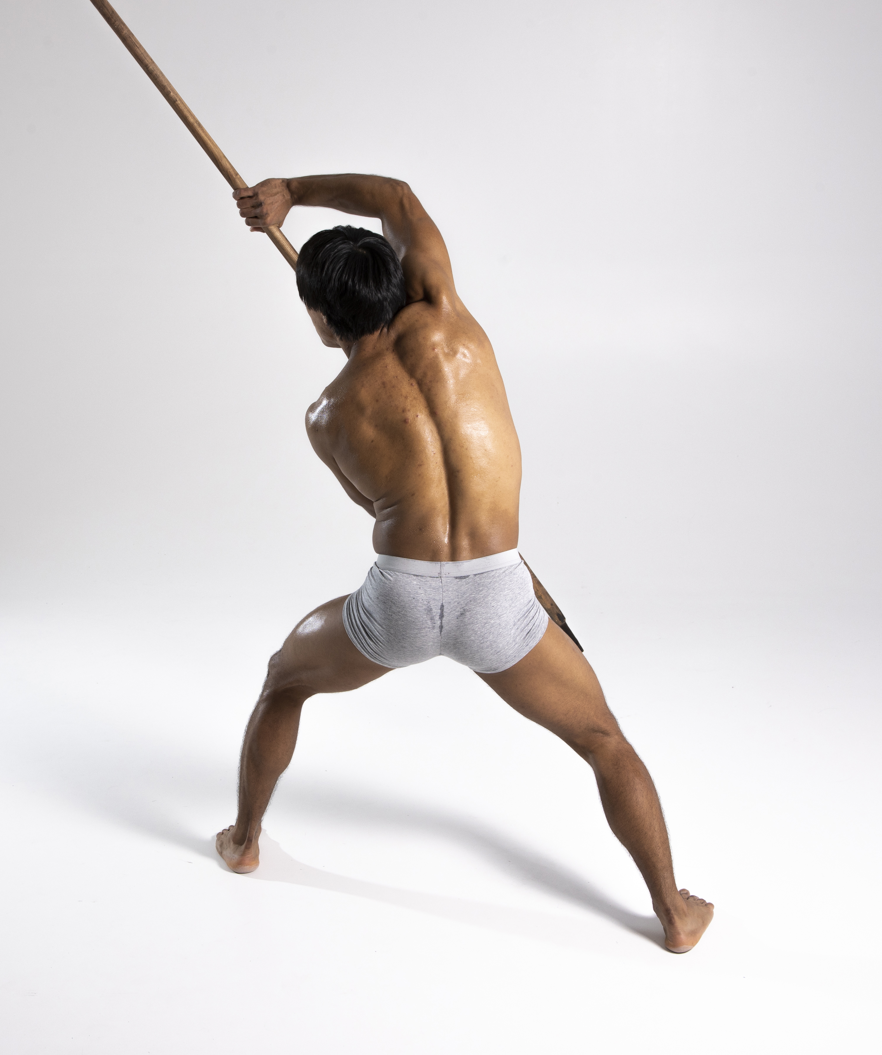a shirtless man in gray underwear is seen from the back, holding a long wooden stick in a fighting stance. he is muscular and athletic. the shot is a studio shot with a white background.