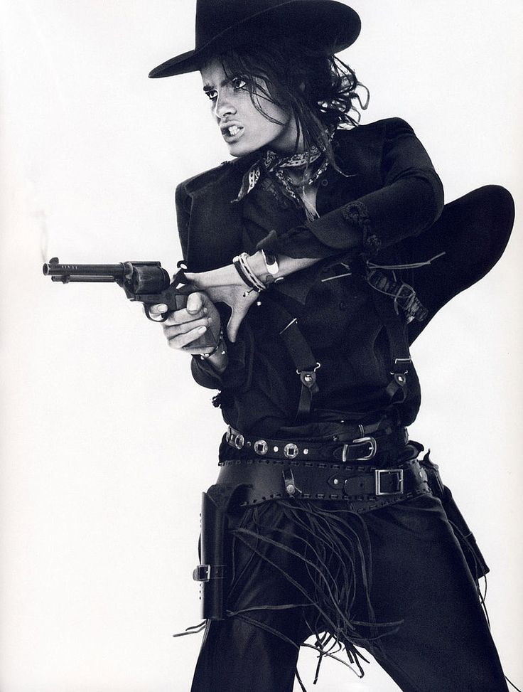 a monochrome portrait of a woman wearing a cowboy hat, black leather vest, belt, and pants. she is holding a gun in her hand and her expression is serious.