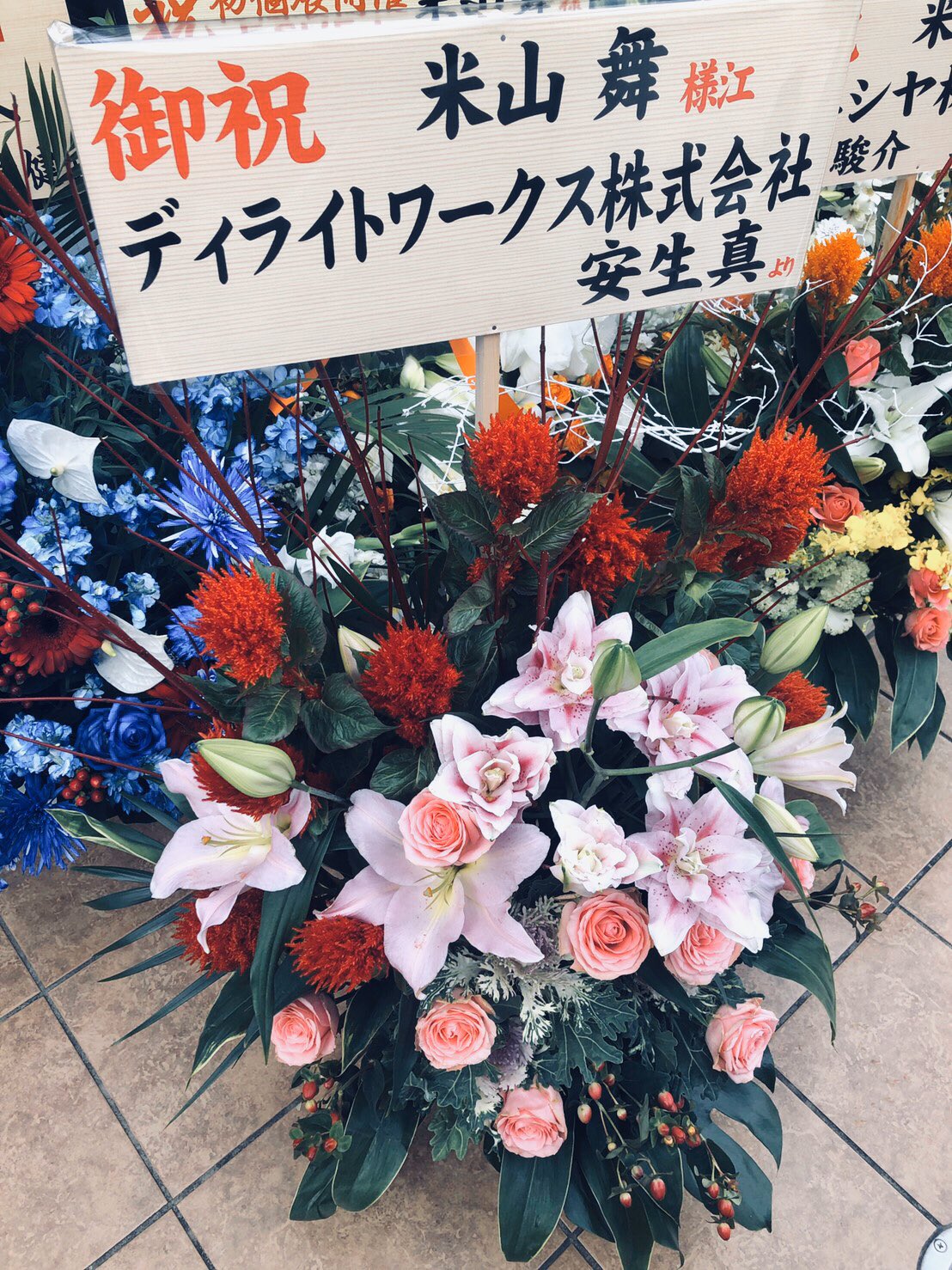 a vibrant floral arrangement, primarily composed of lilies, roses, and carnations in shades of pink, red, and white, sits on a tiled floor. the arrangement is adorned with a sign bearing japanese text, signifying congratulations and likely indicating a grand opening or similar celebratory occasion. the setting appears to be indoors, illuminated by bright, artificial lighting.