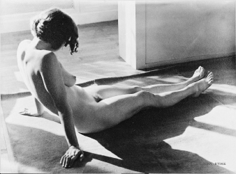 a black and white photograph of a nude woman reclining on a floor. the woman is positioned with her back to the camera, her head turned slightly to the side.  the lighting is dramatic, highlighting the curves of her body and creating strong shadows. the background is simple, with a lightcolored wall and floor. the overall mood is serene and artistic.