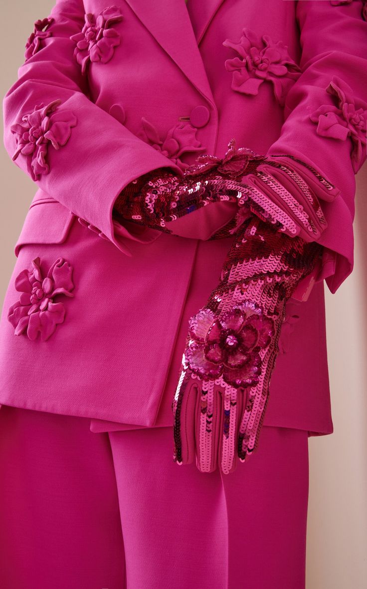loseup shot of a woman wearing a vibrant fuchsia pink suit with intricate floral appliques. the suit consists of a blazer and trousers.  the model is also wearing long sequined gloves with matching floral embellishments. the focus is on the detailed textures and rich color of the clothing. the overall impression is one of highfashion and luxury.