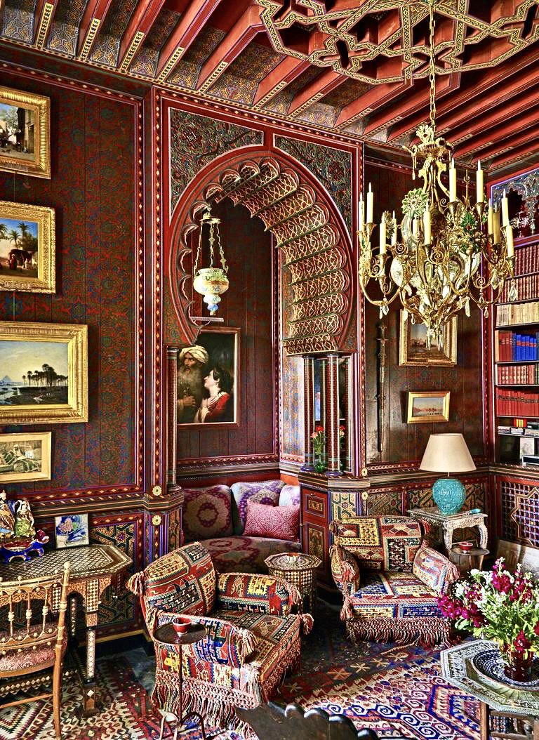 this is a richly decorated room in a moroccan style, featuring ornate patterns, antique furniture, and a mix of vibrant colors like red and gold. the room has a seating area with patterned cushions and tasseled chairs, paintings on the wall, a bookshelf filled with books, and a prominent chandelier hanging from the ceiling. the room also features an archway and a patterned rug on the floor.
